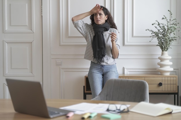 La signora freelance malsana preoccupata controlla il termometro indossando una sciarpa lavorata a maglia che si sente calda dalla febbre