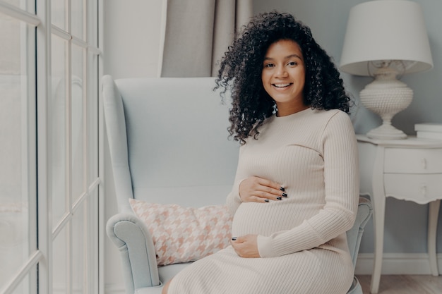 La signora incinta sorridente adorabile afroamericana in abito beige si siede in poltrona bianca