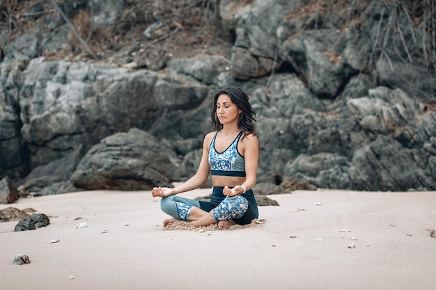La signora snella felice medita nella posa del loto sulla spiaggia sabbiosa vicino alle rocce, concetto di yoga
