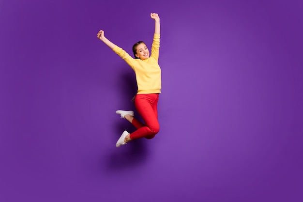 La signora teenager graziosa integrale che salta in alto celebra il tempo di vacanza alzando le mani in alto indossare pantaloni rossi pullover gialli casual parete di colore viola isolato