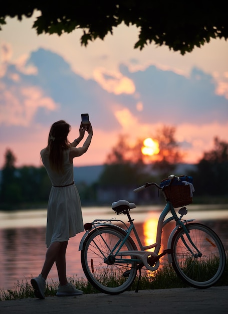 La signora vicino alla bici retrò scatta la foto del tramonto sul cellulare vicino al lago