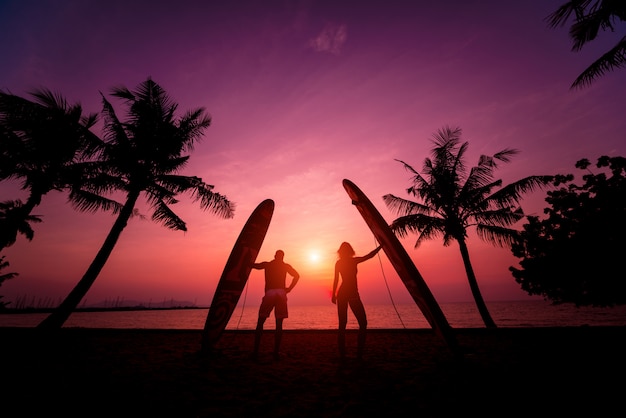 La siluetta dei surfisti coppia la tenuta dei bordi di spuma lunghi al tramonto sulla spiaggia tropicale