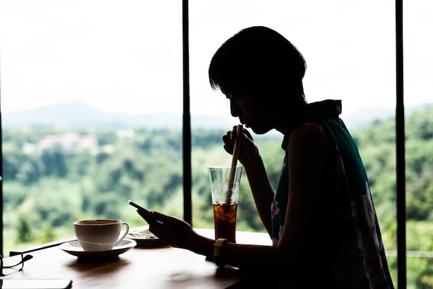 La siluetta della donna asiatica usa il telefono cellulare e prende un drink freddo nella caffetteria