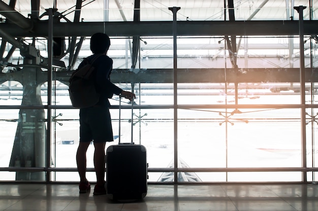 La siluetta di un viaggiatore con la valigia e lo zaino dei bagagli guarda fuori dalla finestra all&#39;aeroporto