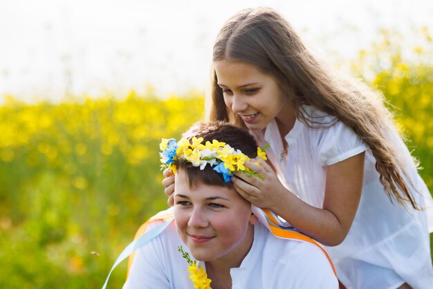 La sorella intreccia i nastri in una corona ucraina con fiori sulla testa del fratello maggiore sul prato contro il campo