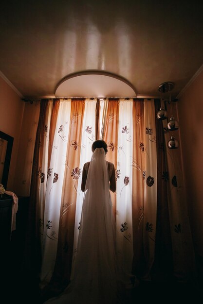 la sposa caucasica in un abito da sposa apre le tende della finestra della camera da letto, foto grandangolare