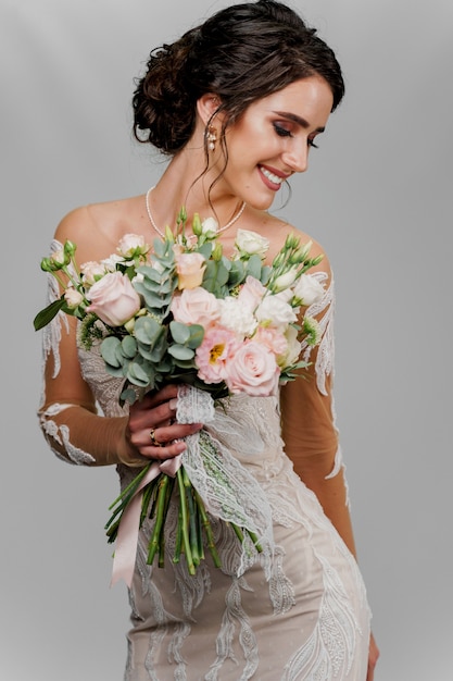 La sposa con il bouquet da sposa guarda a destra e sorride. Ritratto verticale della ragazza attraente per i social network. Ragazza in abito da sposa sul muro bianco.