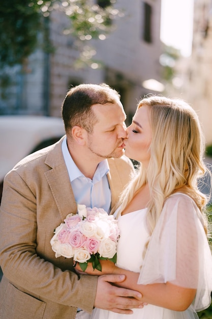 La sposa con il mazzo e lo sposo stanno abbracciando e baciando su un'accogliente strada del primo piano della città vecchia