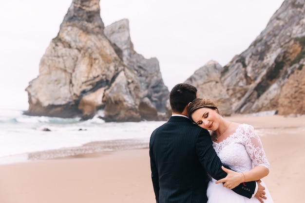 La sposa con un bel trucco si è appoggiata allo sposo sullo sfondo dell'oceano e delle scogliere. avvicinamento. lo sposo torna alla telecamera.