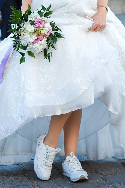 La sposa con un mazzo di cerimonia nuziale si è vestita in un vestito bianco che mostra le scarpe da tennis sulle sue gambe.