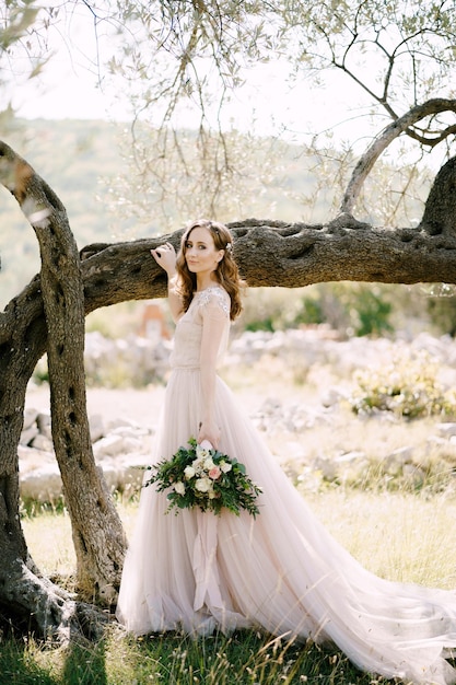 La sposa con un mazzo di fiori sta vicino a un albero con la mano sul tronco