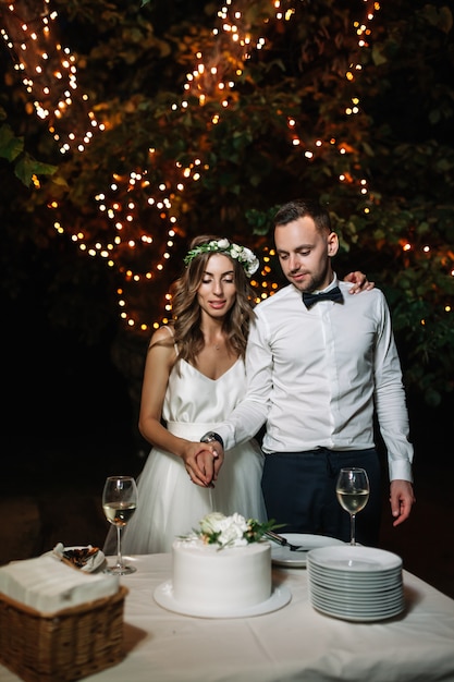 La sposa e lo sposo felici hanno tagliato la torta nuziale nella decorazione leggera anteriore della ghirlanda.