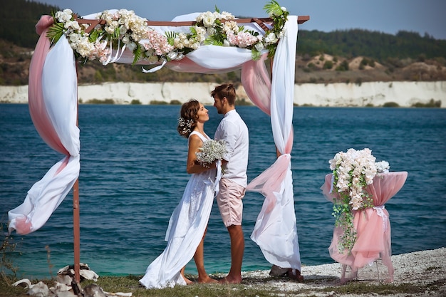 la sposa e lo sposo in abiti bianchi con un mazzo di fiori bianchi stanno sotto un arco di fiori e stoffa sullo sfondo di un lago blu e sabbia bianca