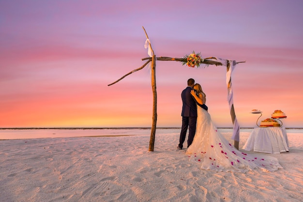 La sposa e lo sposo sotto l'arco sulla spiaggia. Sfondo romantico anniversario di matrimonio di destinazione