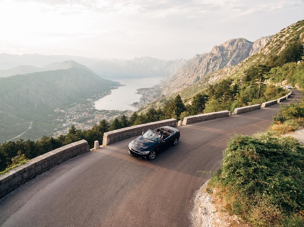 La sposa e lo sposo stanno guidando in una decappottabile su una strada di montagna sullo sfondo della baia di Kotor