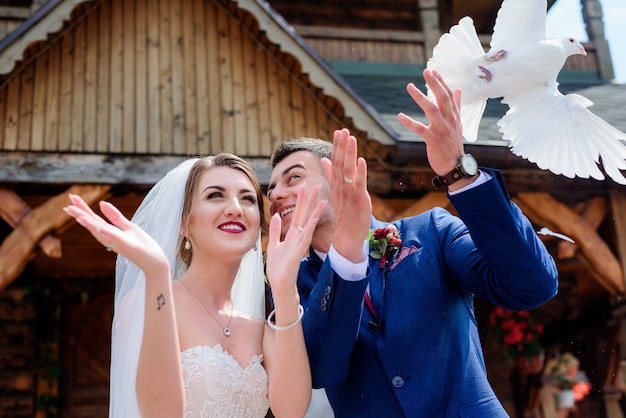 La sposa e lo sposo tengono i piccioni in piedi davanti a una chiesa di legno