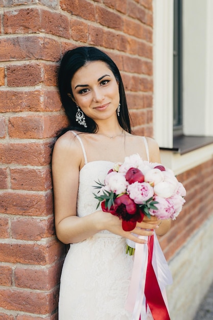 La sposa femminile attraente indossa l'abito bianco da sposa tiene il bouquet vicino al muro di mattoni ha un aspetto accattivante La giovane donna elegante con il trucco ha un sorriso affascinante celebra il suo matrimonio