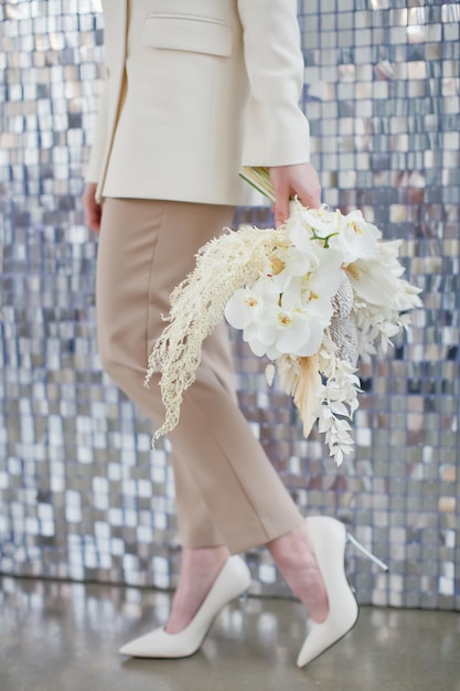 La sposa in abito beige tiene il bouquet da sposa boho con orchidee banksia e fiori secchi su uno sfondo di paillettes argento