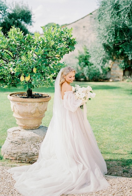 La sposa in abito bianco con un mazzo di rose si trova vicino a un albero in una vasca in giardino