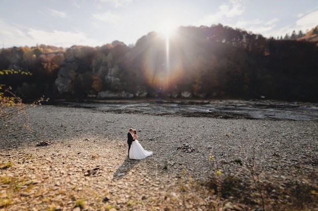 La sposa in abito bianco e lo sposo in piedi vicino al fiume e abbracci e sole