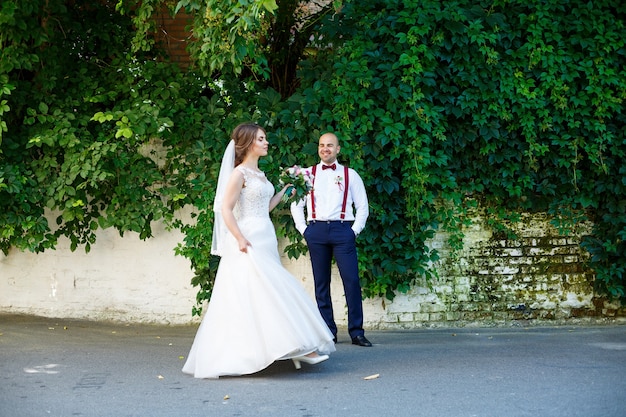 La sposa in abito bianco in primo piano e lo sposo con le bretelle dietro. sul muro di fondo con foglie verdi