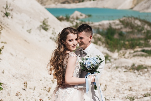 La sposa in un bel vestito che abbraccia lo sposo in una tuta leggera vicino al lago. Coppie di cerimonia nuziale che si levano in piedi su una collina sabbiosa all'aperto. Una romantica storia d'amore. Acqua azzurro-azzurro all'orizzonte.