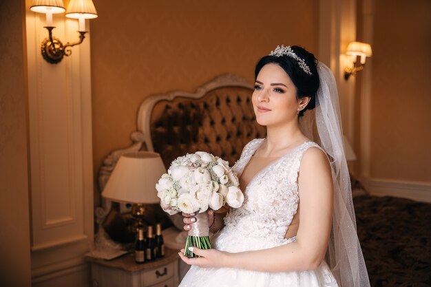 La sposa in un bellissimo abito da sposa tiene un bouquet da sposa e guarda nella telecamera. Sposa su uno sfondo di un'accogliente camera da letto.