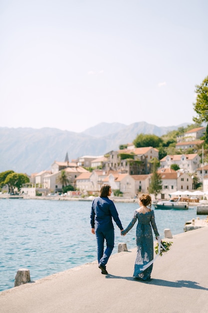 La sposa in un elegante abito blu e lo sposo camminano lungo il molo tenendosi per mano vicino alla città vecchia di Perast, vista posteriore. Foto di alta qualità