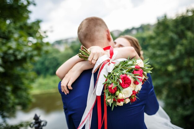 La sposa mette le mani sulle spalle dello sposo sposa con un bouquet di rose rosse e bianche abbraccia e bacia lo sposo in natura