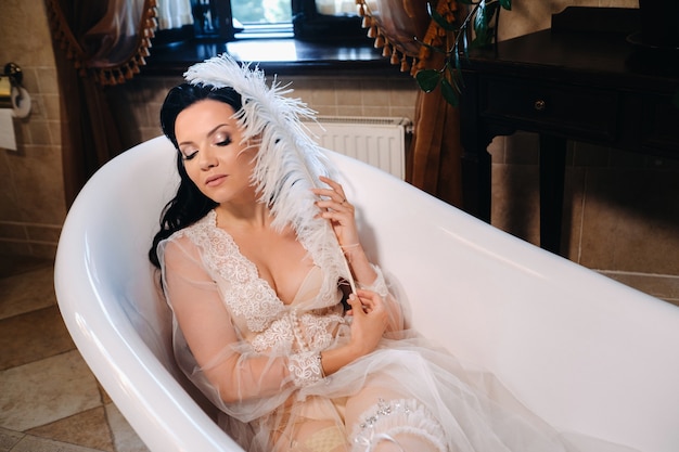 La sposa, vestita con un abito trasparente da boudoir e biancheria intima, giace in un bagno vintage con una piuma bianca tra le mani, La mattina della sposa