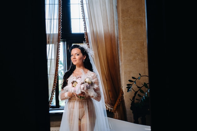 La sposa, vestita con un abito trasparente da boudoir e biancheria intima, si siede vicino a un bagno vintage con una piuma tra le mani, La mattina della sposa.