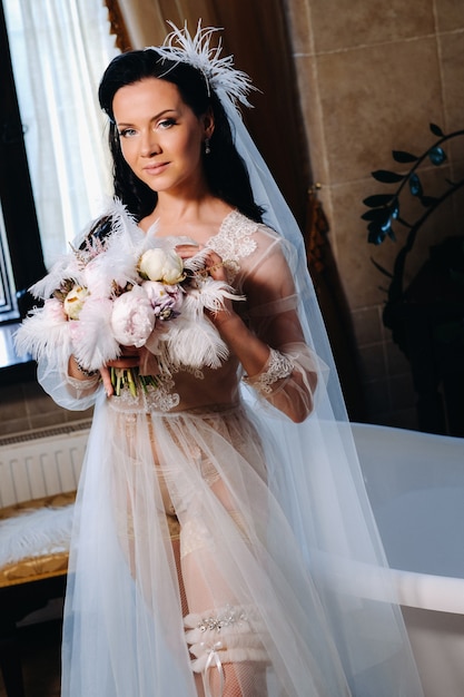 La sposa, vestita con un abito trasparente da boudoir e biancheria intima, si siede vicino a un bagno vintage con una piuma tra le mani, La mattina della sposa.