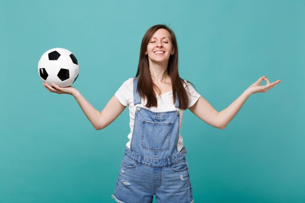 La squadra preferita di sostegno del tifoso di calcio della donna sorridente con il pallone da calcio si tiene per mano nel gesto di yoga, meditando rilassante isolato su sfondo blu turchese. Emozioni della gente, concetto di svago della famiglia di sport.