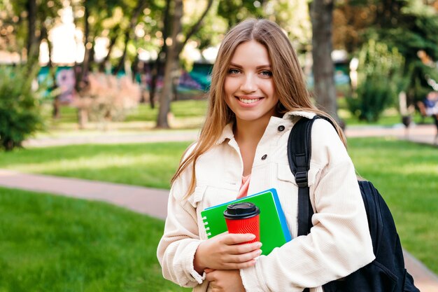 La studentessa bionda cammina nel parco con un taccuino e una tazza di caffè, spazio della copia. Concetto di educazione