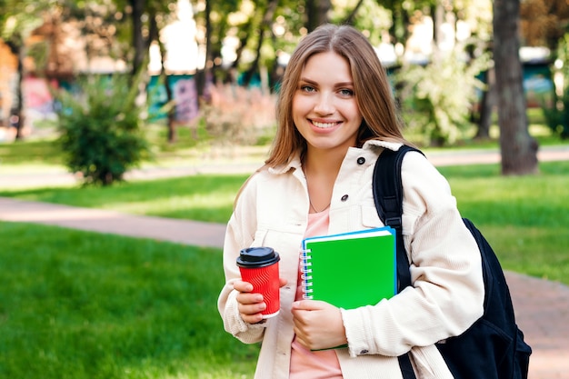 La studentessa bionda cammina nel parco con un taccuino e una tazza di caffè