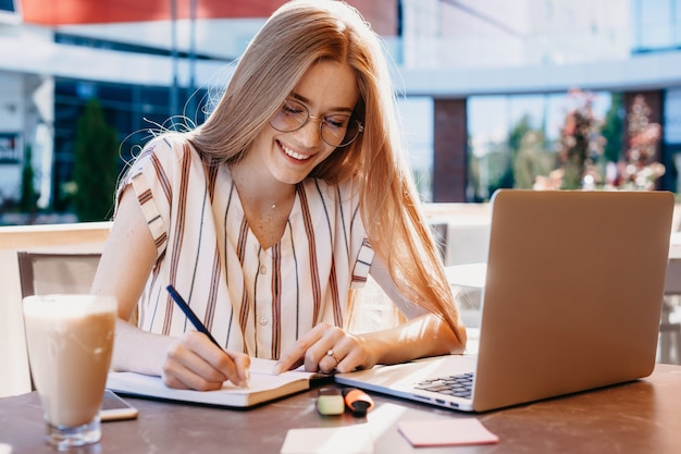 La studentessa caucasica allo zenzero con le lentiggini sta scrivendo qualcosa sul suo taccuino mentre beve un caffè e usa il suo laptop