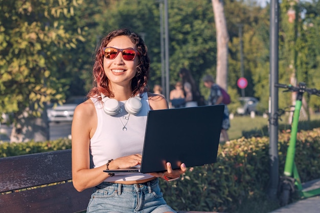 La studentessa con le cuffie sta lavorando e studiando su un laptop seduto sulla panchina nel parco