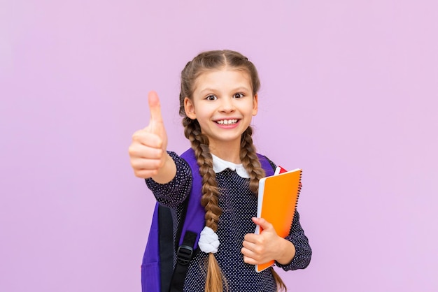 La studentessa dà un pollice in alto e sorride ampiamente Una bambina con uno zaino e quaderni per studiare su uno sfondo rosa isolato