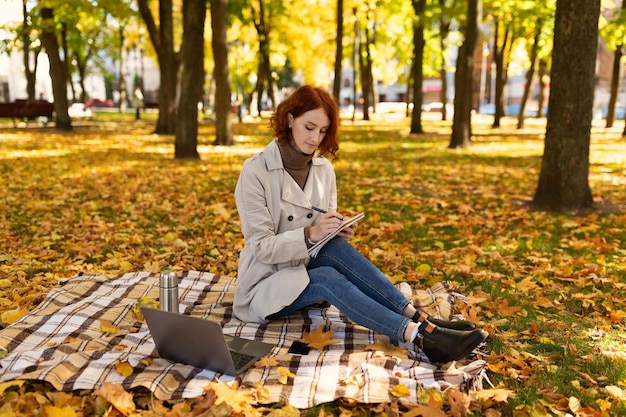 La studentessa millenaria caucasica occupata intelligente pensierosa con capelli rossi in impermeabile si siede sul plaid con il libro e il computer portatile