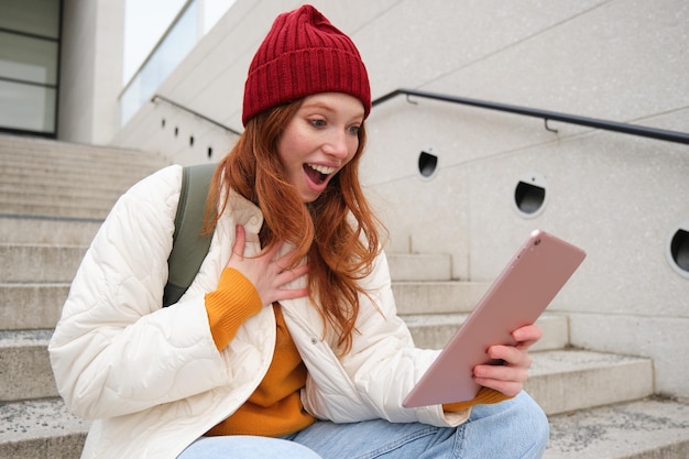 La studentessa rossa alla moda felice con il cappello rosso tiene il tablet digitale utilizza le ricerche di app sui social media così