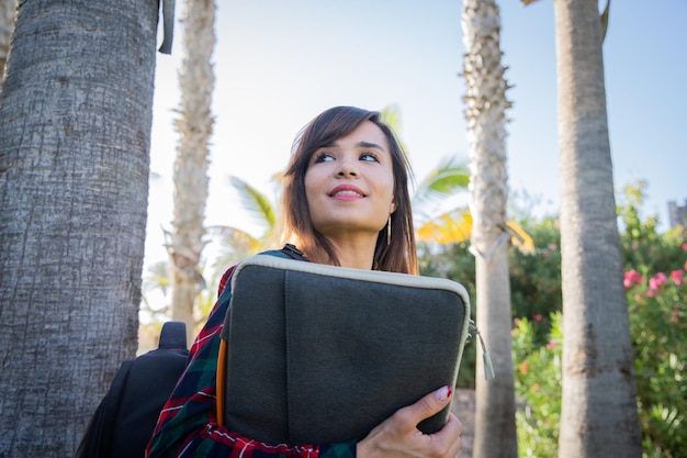 La studentessa universitaria distoglie lo sguardo mentre tiene in mano la custodia del laptop e indossa lo zaino