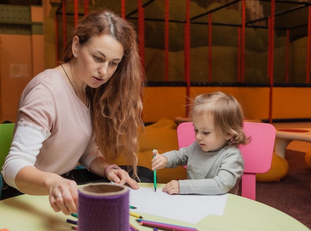 La tata insegna a una bambina come disegnare con le matite colorate mentre è seduta a un tavolo in un centro di intrattenimento per bambini