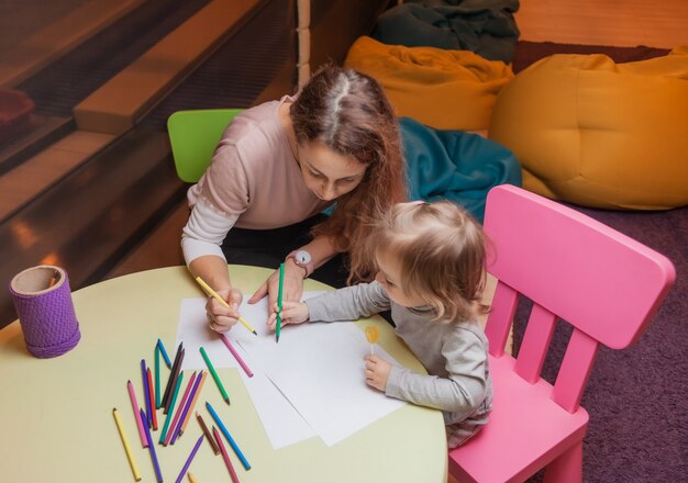 La tata insegna a una bambina come disegnare con le matite colorate mentre è seduta a un tavolo in un centro di intrattenimento per bambini