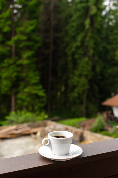 La tazza bianca di caffè mattutino caldo scuro su recinzione di legno con sfondo di foresta.