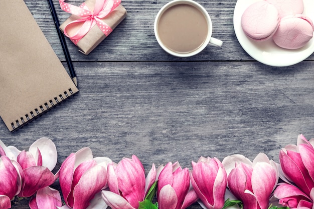 La tazza di caffè di mattina con latte, macaron del dolce, il regalo o la scatola attuale e la magnolia fiorisce sulla tavola di legno rustica. disteso