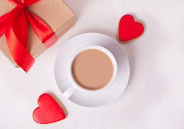 La tazza di caffè e un cuore hanno modellato i biscotti rossi con il contenitore di regalo sulla tavola bianca