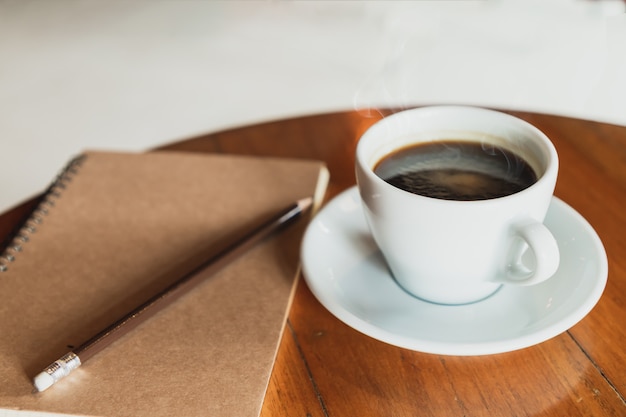 La tazza di caffè macchiato del primo piano, la matita, taccuino marrone della copertura ha messo sopra una tavola di legno marrone in una caffetteria