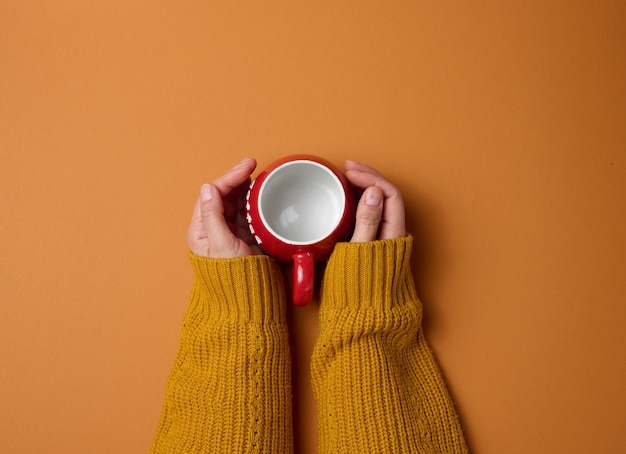La tazza di ceramica rossa vuota in una mano femminile su sfondo arancione, la bevanda e la mano sono sollevate, pausa caffè