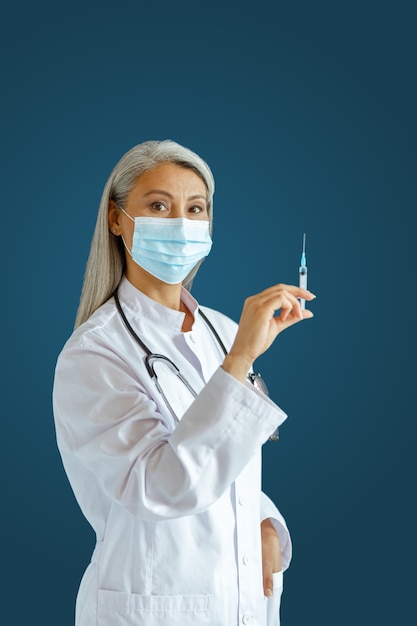 La terapista asiatica dai capelli d'argento in uniforme con maschera protettiva tiene la siringa con la medicina in piedi su sfondo blu in studio. Giornata della vaccinazione