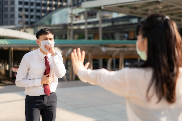 La tosse asiatica dell'uomo d'affari malato con maschera e segnale di stop della donna di affari lo passa per proteggere la distanza dai virus COVID-19 e dalle persone che prendono le distanze sociali per il rischio di infezione